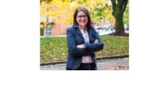 color photo of Carli Friedman, a young white woman with straight brown hair and glasses wearing a blue blazer and white shirt. She is standing with her arms crossed and is smiling. She is outside on what looks like a college campus.