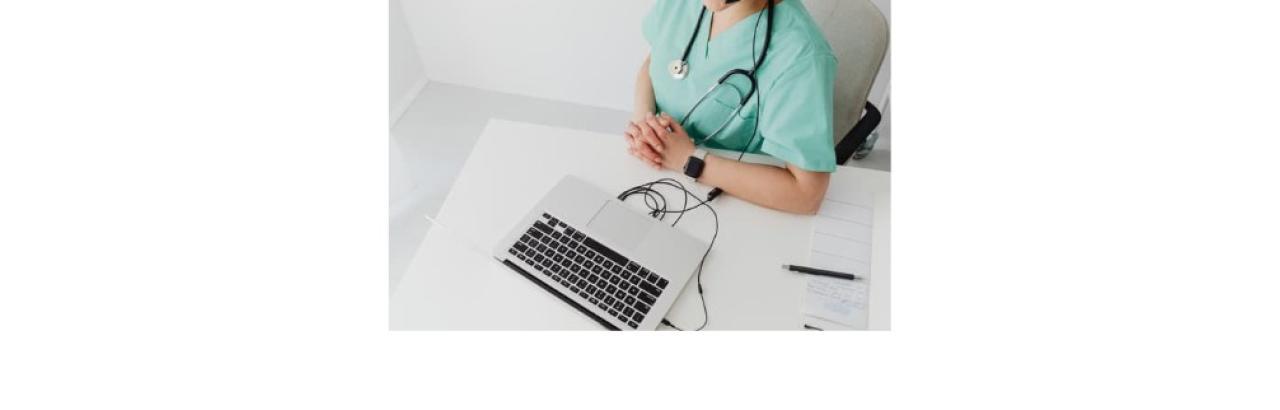 Photo of a person wearing blue scrubs sitting at a table in front of a laptop. Their head isn't shown in the picture. The person is white and has a stethoscope around their neck and a smart watch